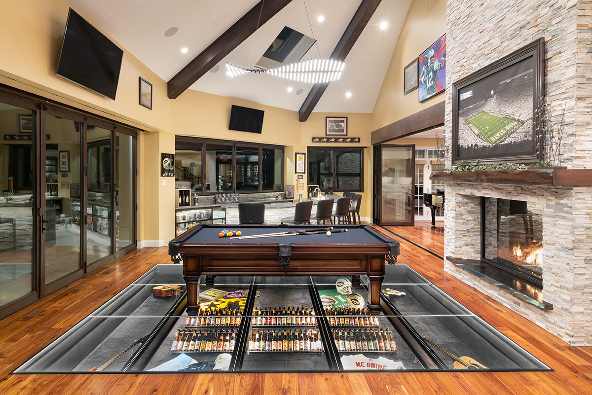 A living room with an interior glass floor in the center of the room displaying sports memorabilia under the glass with a pool table sitting on top of the glass floor.