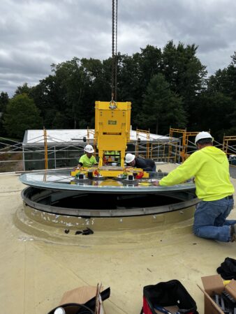 Workers securing a circular glass skylight on a roof, ensuring stability and precision in installation against a backdrop of construction scaffolding.