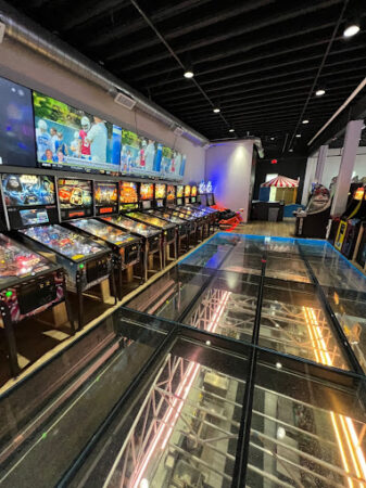 A row of pinball machines next to a transparent glass floor that reveals structural beams below, creating a dynamic, multi-layered look in the arcade.