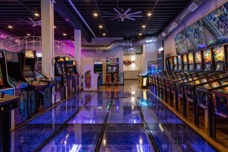 A vibrant arcade with retro gaming machines lining the walls. The centerpiece is a reflective glass floor with blue lighting, creating a lively atmosphere.