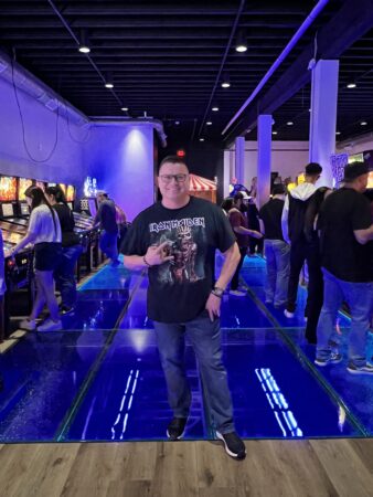 A man stands on an illuminated glass floor in an arcade, surrounded by people playing pinball machines. The floor glows in blue, enhancing the retro vibe.