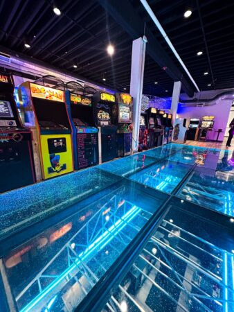 Close-up of a glowing blue glass floor with retro arcade games, creating a futuristic yet nostalgic ambiance under colorful neon lights.