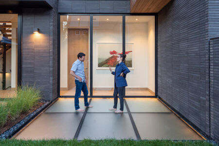 Two individuals stand on a translucent glass floor, discussing architectural design in front of a sleek, modern building with expansive glass windows.