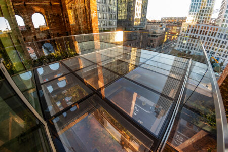 Close-up of a glass deck terrace reflecting the golden hues of the sunset with views of modern high-rises and historic brick architecture in the background.