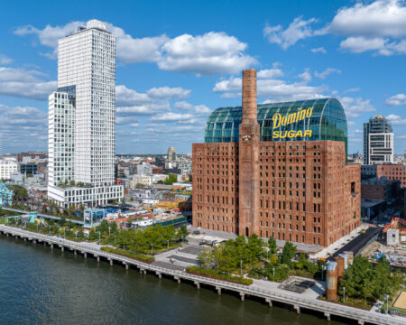 Aerial view of the historic Domino Sugar Refinery building with its iconic sign, surrounded by modern high-rise buildings and waterfront parkland.