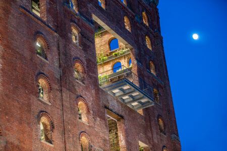 The historic brick facade of Domino Refinery featuring a stunning cantilevered glass balcony illuminated against the evening sky with a full moon.