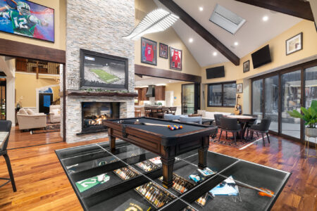 Spacious modern living room with vaulted ceilings, a fireplace, and a glass floor showcasing memorabilia under a pool table, merging luxury and creativity.