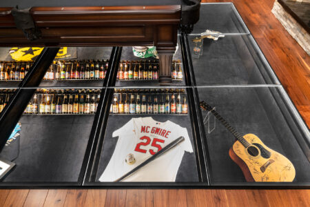 A glass floor beneath a pool table reveals signed jerseys, guitars, and beer bottles, combining functionality with a stunning display of cherished items.