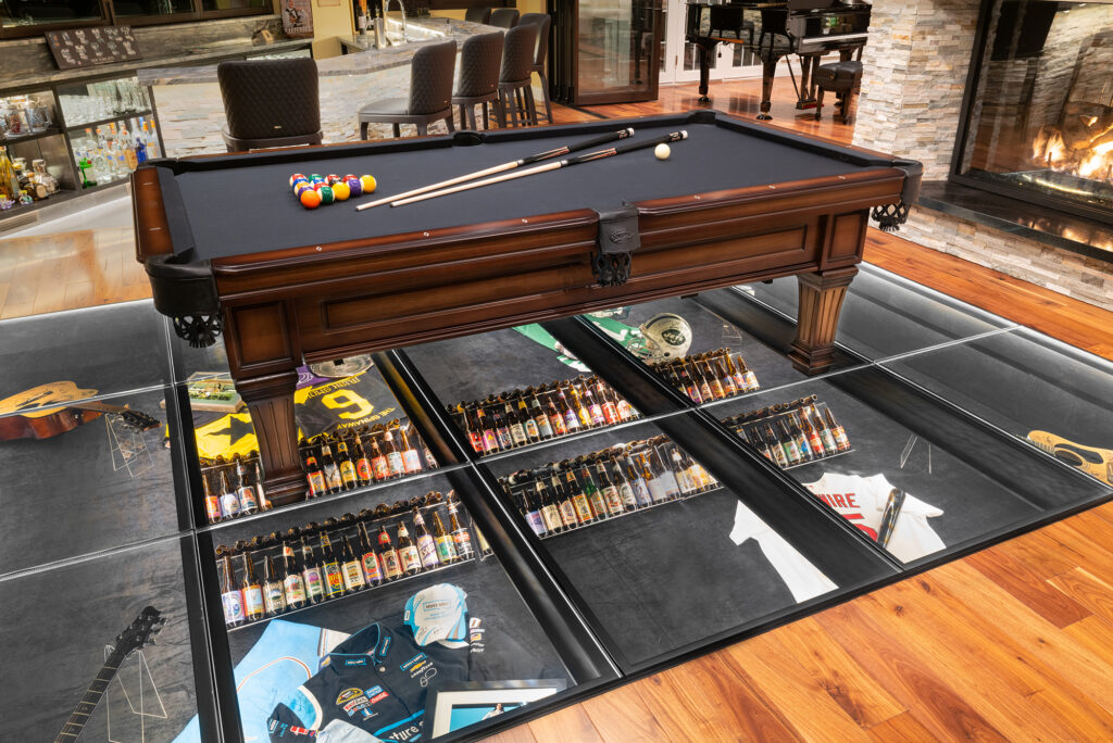 Elegant pool table centered on a glass floor, displaying organized memorabilia below, blending entertainment and innovative design in a luxury space.