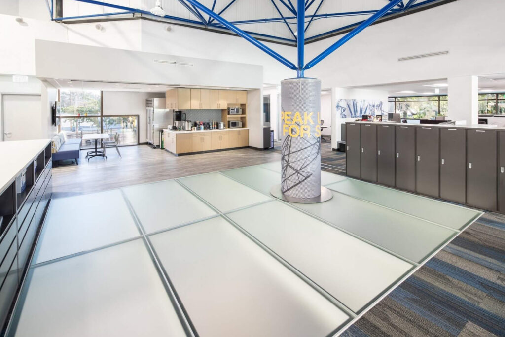 A wide-angle view of a modern office featuring frosted glass flooring, vibrant blue beams, and an open, collaborative environment.