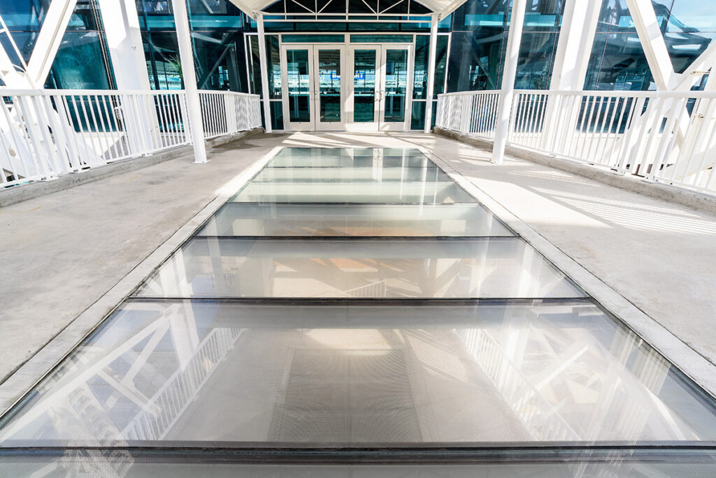 A modern glass walkway with anti-slip textured panels, leading to a building entrance with white metal railings on both sides.