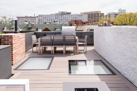 Modern rooftop deck with two walkable glass skylights, an outdoor dining area, and built-in grill, overlooking an urban cityscape.