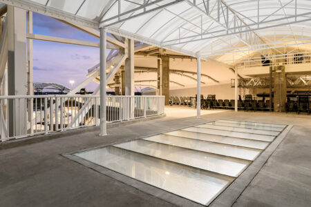 A spacious covered outdoor area with a glass floor installation, framed by white structural beams and overlooking a bridge.