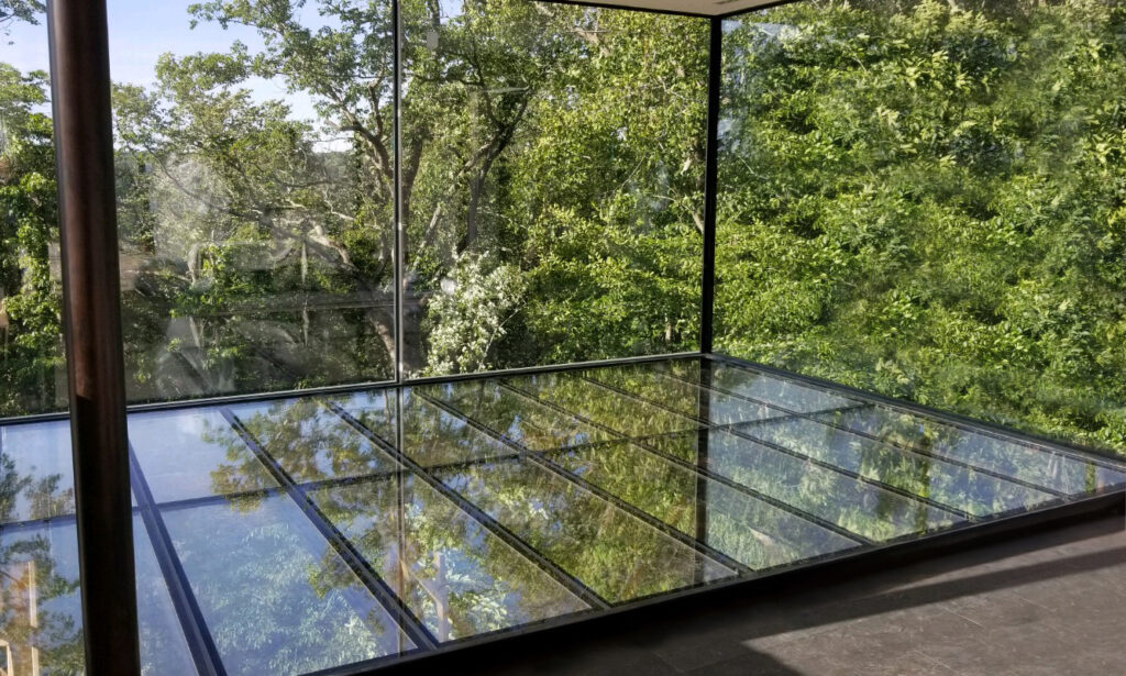 Two individuals stand on a translucent glass floor, discussing architectural design in front of a sleek, modern building with expansive glass windows.