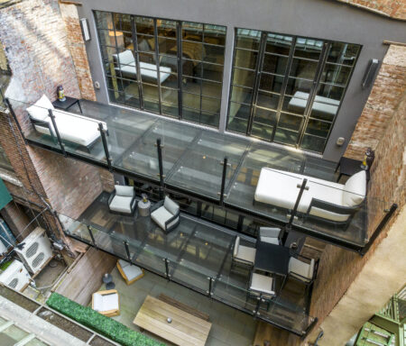 Stacked glass balconies with black-framed guardrails extend from a brick apartment complex overlooking a courtyard.