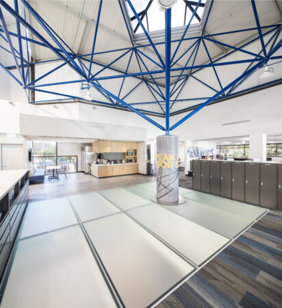 Bright open-concept office space featuring frosted glass flooring around a central blue steel column under a skylight.