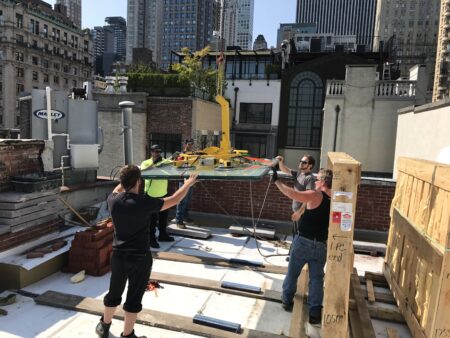 Contractors installing glass panel on rooftop using crane system with New York City skyline in background.
