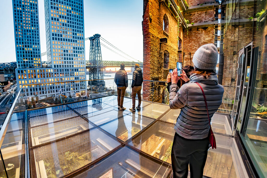 Tourists on a glass terrace take in the Manhattan skyline and Williamsburg Bridge at sunset at The Refinery, NYC.