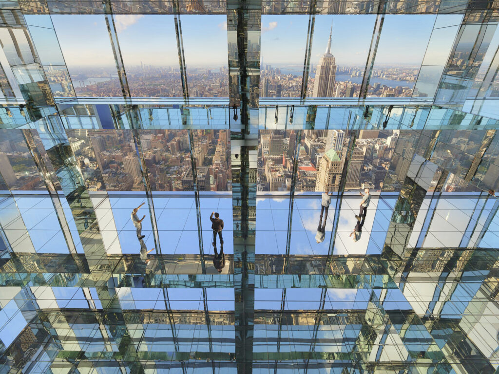 Visitors walk across transparent glass floors in a high-rise building with panoramic views of New York City.