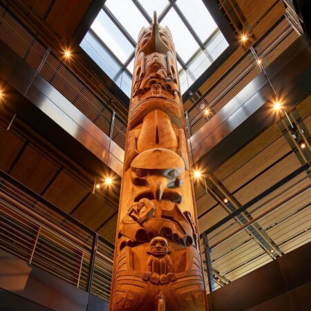 A towering carved totem pole beneath a structural skylight in a wood and steel interior with track lighting above.