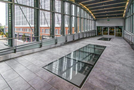 A spacious commercial walkway with framed glass floor insets, surrounded by floor-to-ceiling windows and steel support beams.