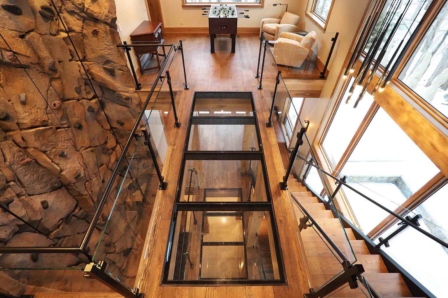 A clear glass walkway with black framing connects two loft areas in a wood-accented home with floor-to-ceiling windows.