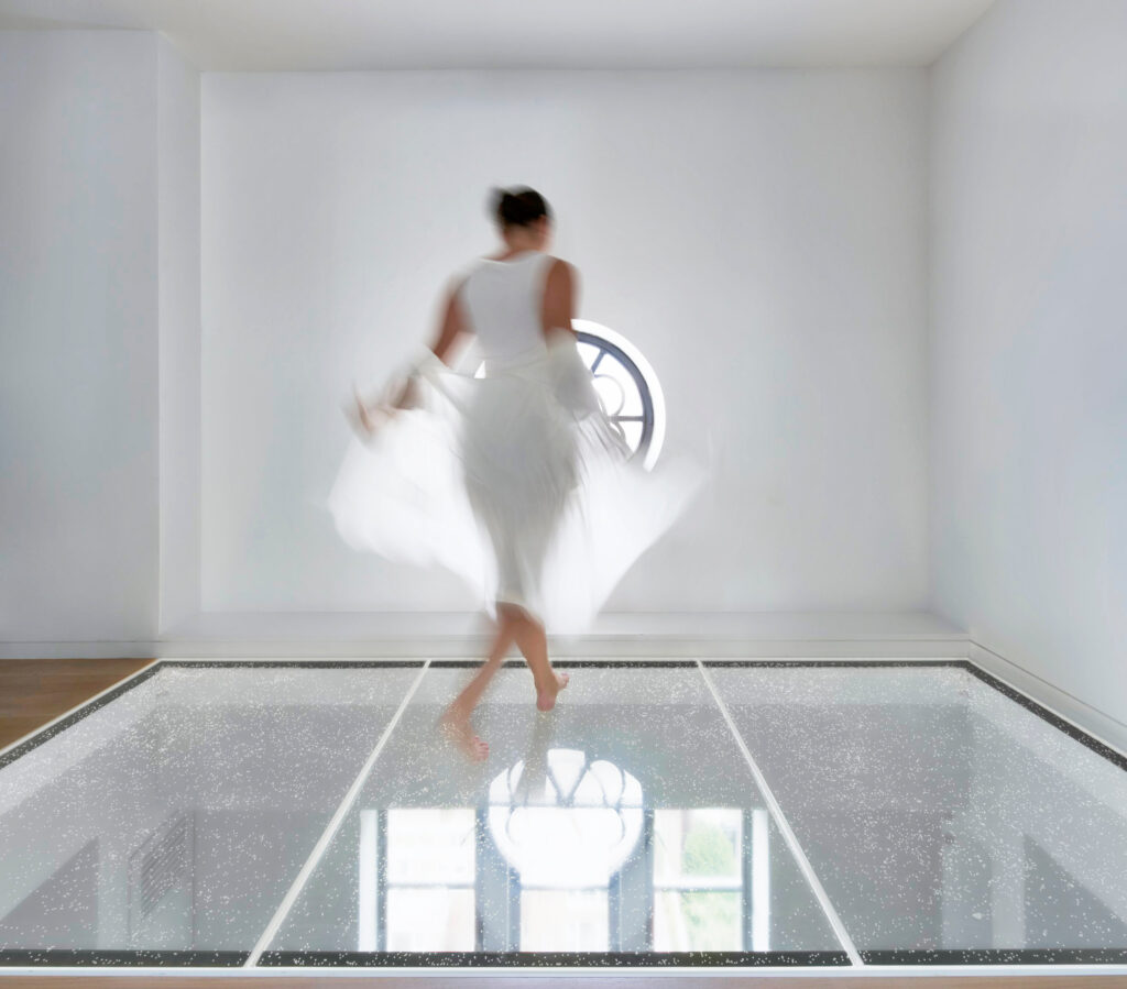 A woman in a white dress twirls on a sparkly glass floor in a minimalistic white room with a circular window in the background.