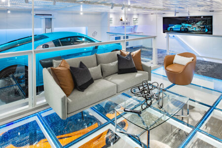 A high-end living space with a gray couch and glass coffee table is built above a glass floor, showcasing a bright blue luxury sports car in the garage below.