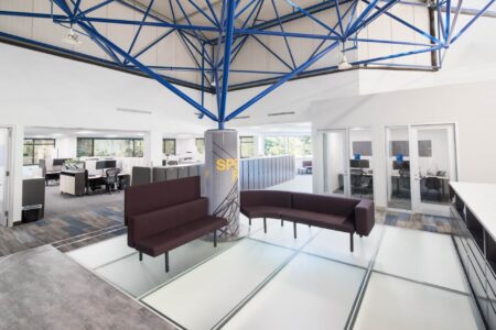 A frosted glass floor section in a bright, open-concept office with blue ceiling trusses and maroon seating around a central column.
