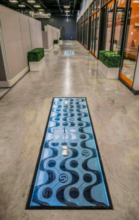 A modern office hallway featuring decorative glass floor panels with a blue and black wavy design, set in polished concrete flooring.