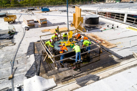 A team of construction workers in safety gear installing a large circular glass skylight on a commercial rooftop under bright daylight.