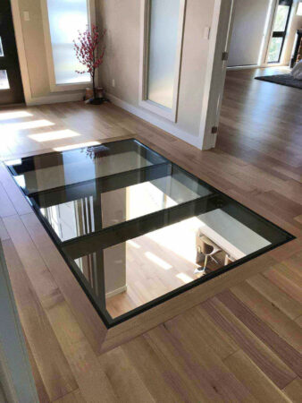 A simple, modern hallway with light wood flooring features a rectangular interior glass floor panel offering a view into the workspace below.