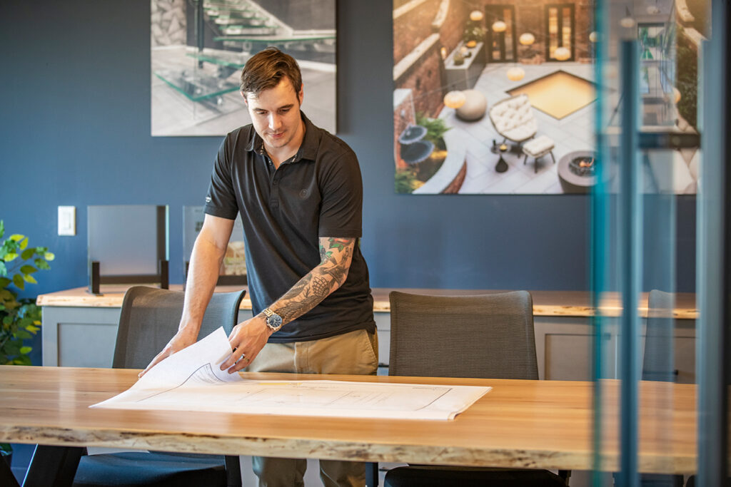 A man reviewing architectural plans in a modern meeting room with mounted glass flooring photos and material samples behind him.