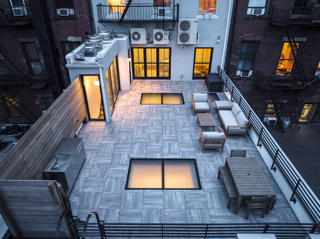 A wide view of a furnished rooftop terrace showcases three glowing glass skylights embedded in the floor, adding both style and natural light access.
