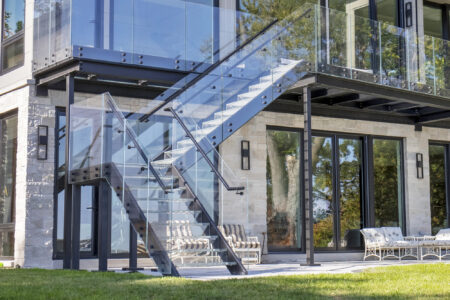 An exterior glass staircase leads to a balcony, with clear railings and metal framing attached to a modern stone-and-glass residential facade.