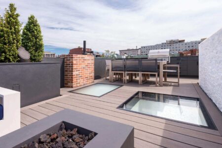 A modern rooftop deck features two walkable skylights set into a composite wood deck, surrounded by outdoor dining furniture and a built-in grill.