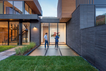 Two people stand on a frosted glass walkway set between sections of a contemporary home, surrounded by black siding and large glass windows.