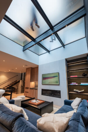 An interior view of a living space reveals frosted glass panels above, with silhouettes of feet walking across a structural glass skylight above.