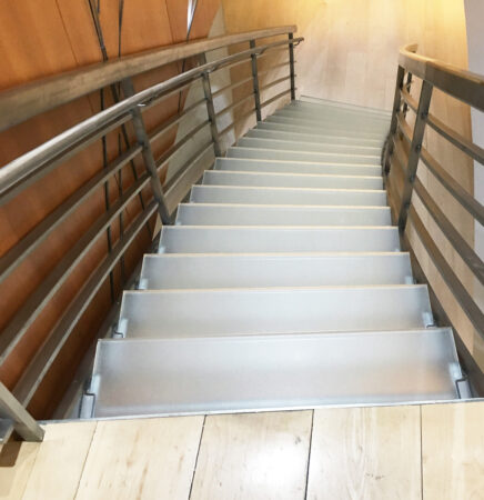 A modern glass staircase with frosted textured treads is flanked by sleek metal railings and light wood-paneled walls.