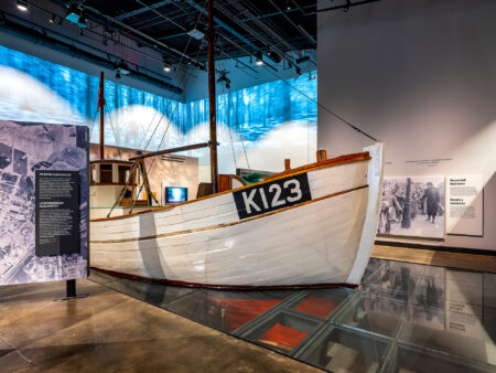 A wooden boat displayed in a museum exhibit resting on a transparent glass floor that reveals artifacts beneath the surface.