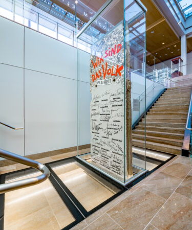 A detailed view of a Berlin Wall segment enclosed in a glass case at the National Museum of American Diplomacy, adjacent to stairs in a sleek, modern interior.