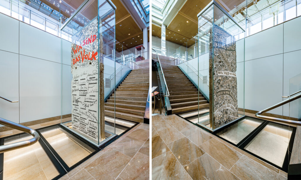 A preserved segment of the Berlin Wall displayed in a tall glass case near a staircase at the National Museum of American Diplomacy, surrounded by a modern architectural design.