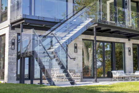 An exterior glass staircase leads to a balcony, with clear railings and metal framing attached to a modern stone-and-glass residential facade.