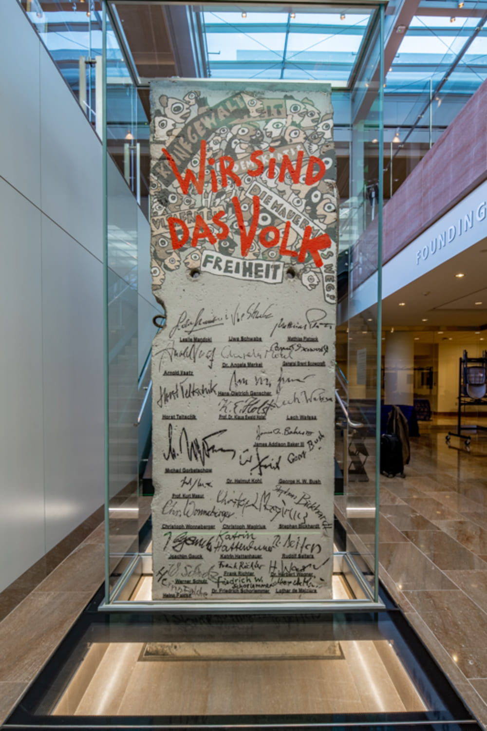Front view of a vertical glass memorial panel with handwritten signatures and red lettering, positioned on an illuminated glass platform indoors.