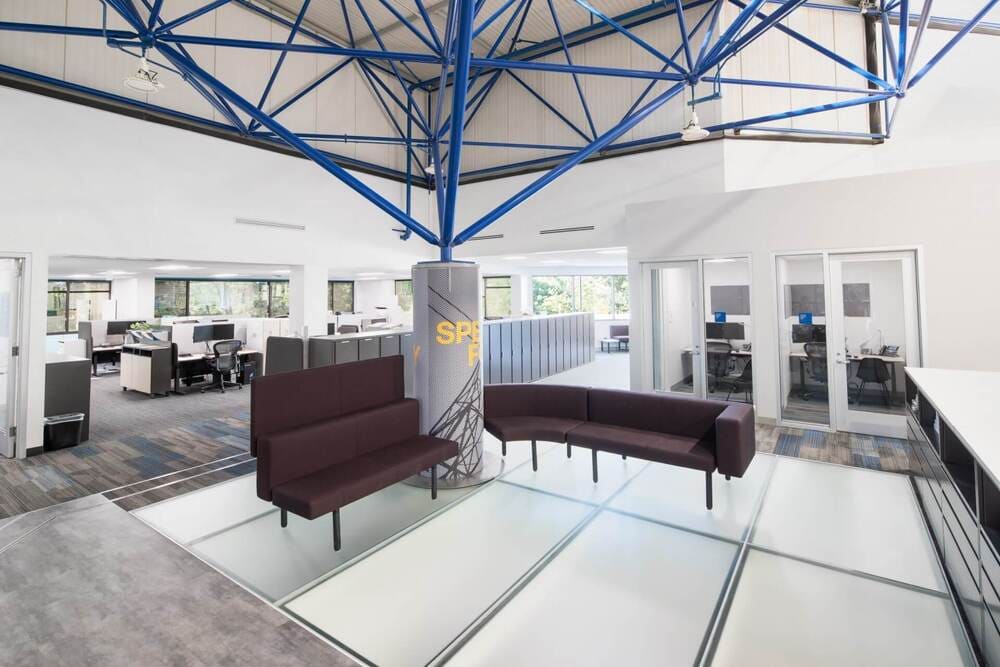 Modern lounge area with sofas placed on a glass floor, blue steel ceiling structure above.