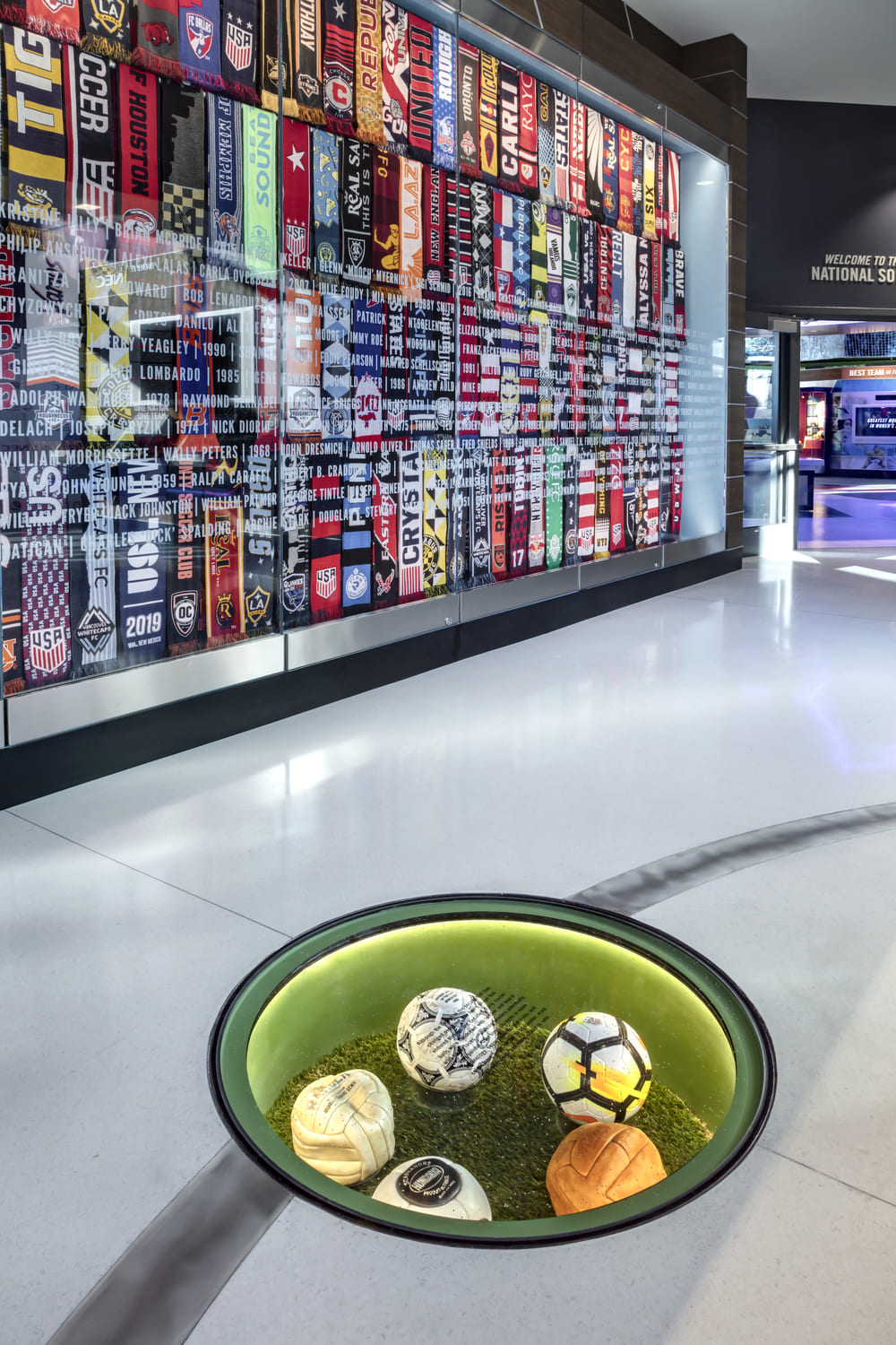 Round glass floor display containing signed soccer balls on artificial grass.