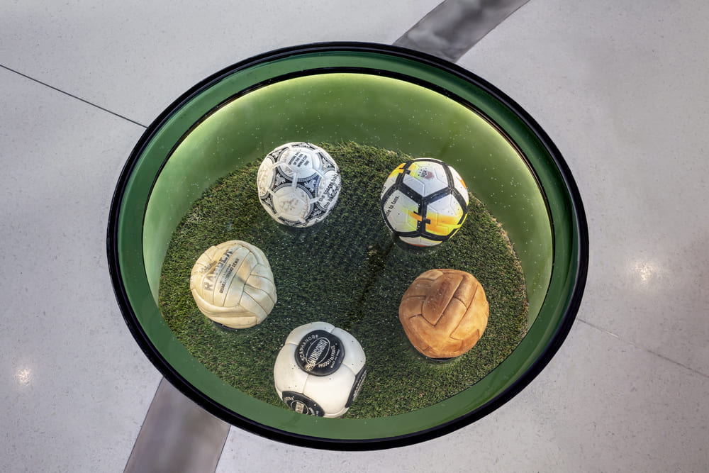 Round glass floor display containing signed soccer balls on artificial grass.