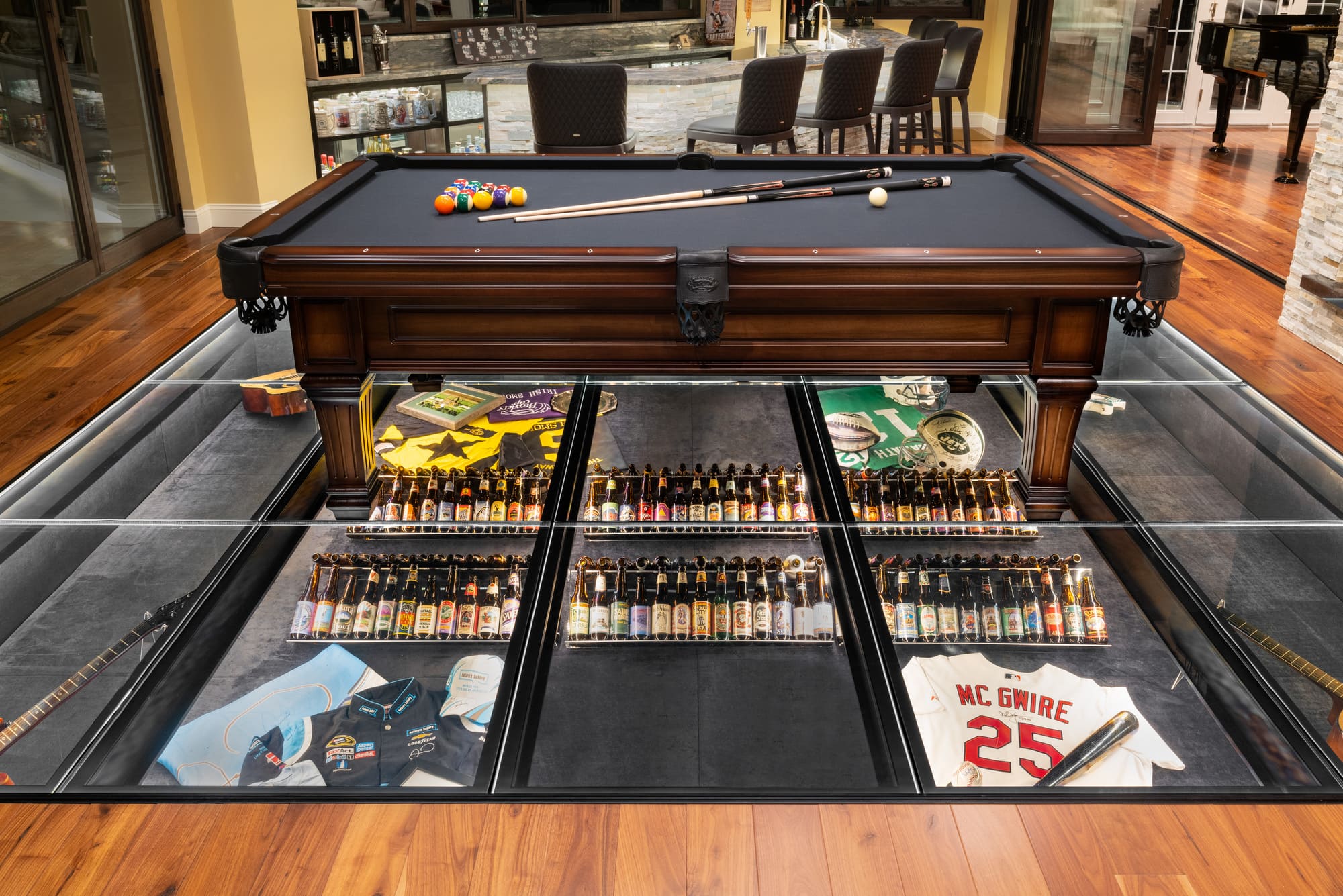 A pool table situated on top of a collection of beer bottles, baseball and football uniforms, and guitars, which are encased in tempered glass.