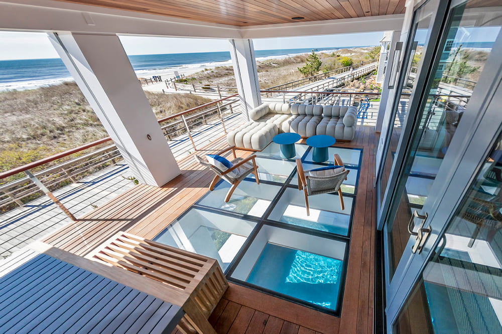 A patio of a modern beach house with a glass panel floor that shows the pool underneath, and an overlooking view of the beach.