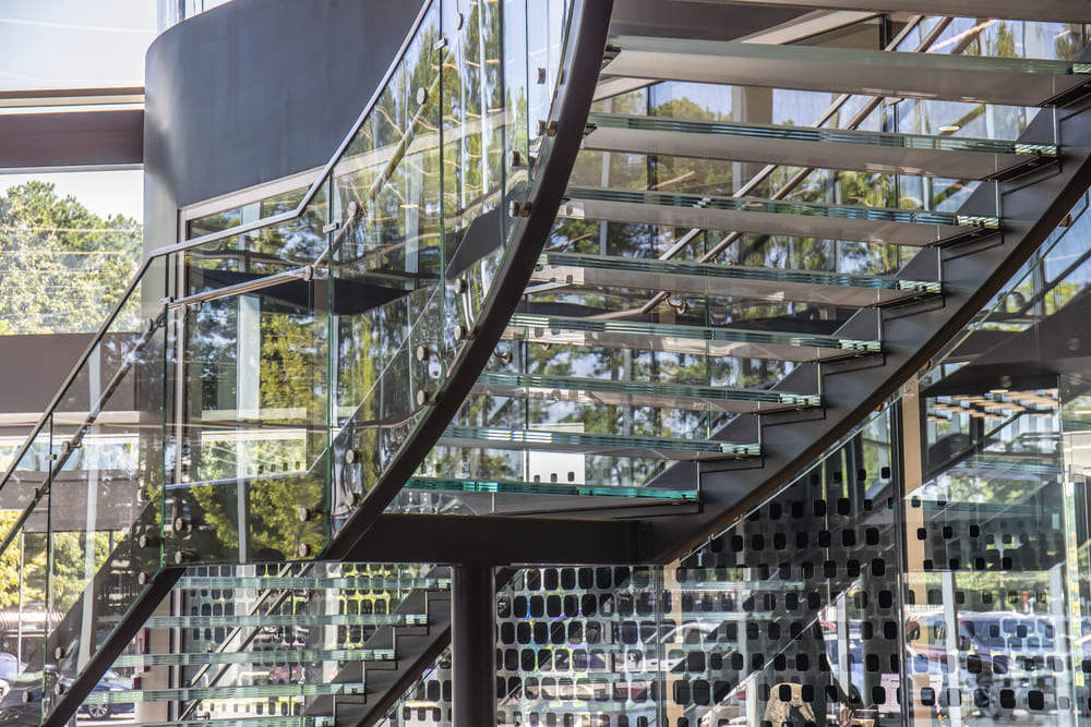 Curved glass staircase in a sunlit modern lobby with reflective glass walls.